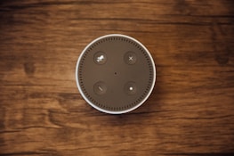 A top-down view of a circular smart speaker with buttons on its surface, placed on a wooden background.