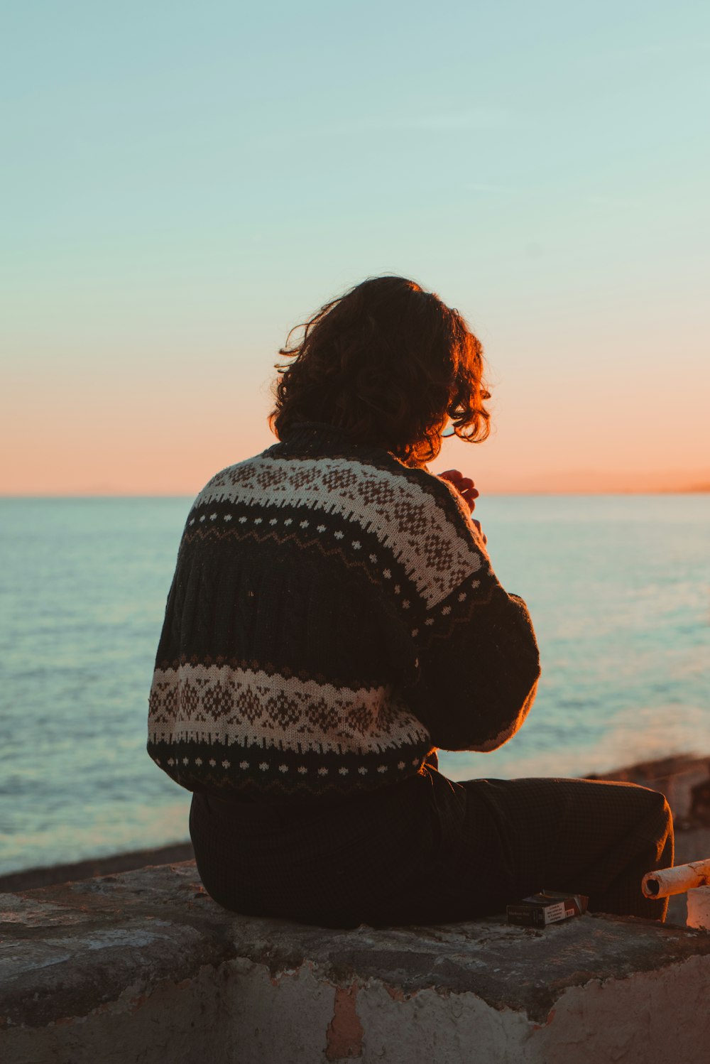 person in black and white sweater sitting on beach shore during sunset