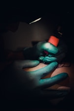 A tattoo artist, wearing teal gloves, is working on a tattoo on a person's skin. The lighting is dim, focusing on the artist's hands holding a tattoo machine with a red grip.