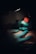 A tattoo artist, wearing teal gloves, is working on a tattoo on a person's skin. The lighting is dim, focusing on the artist's hands holding a tattoo machine with a red grip.