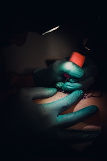A tattoo artist, wearing teal gloves, is working on a tattoo on a person's skin. The lighting is dim, focusing on the artist's hands holding a tattoo machine with a red grip.
