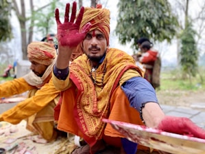 A man wearing traditional attire with a turban and a colorful shawl is holding out his hand, which is covered in red powder. Another person dressed similarly is seated beside him. In the background, there are trees and a blurred figure.