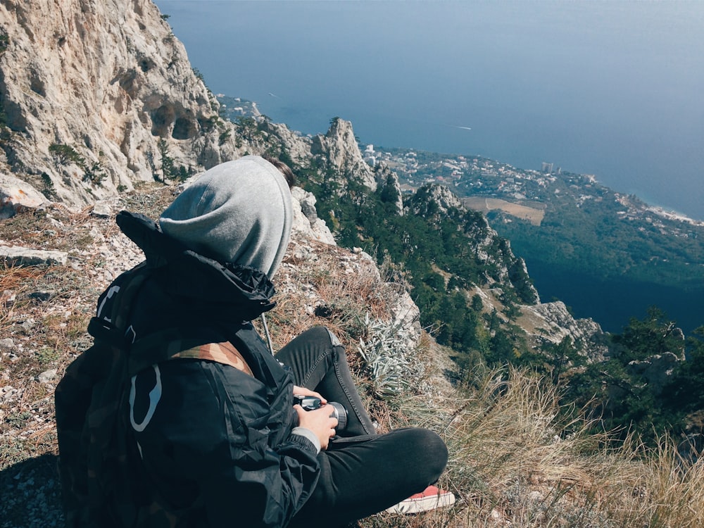 personne en veste noire et bonnet en tricot gris assis sur rock mountain pendant la journée