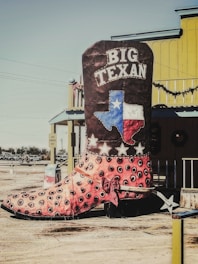 A large decorative cowboy boot with the text 'Big Texan' is displayed outdoors. The boot features a design with eye-catching colors and patterns, including a depiction of the Texas flag and stars. The setting includes a bright yellow building with western-style architectural elements in the background.