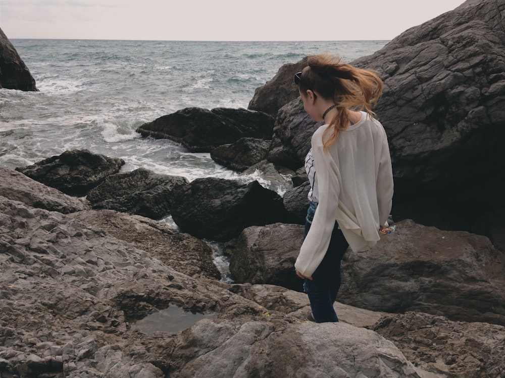 Frau in weißem Langarmhemd steht tagsüber auf einer Felsformation in der Nähe des Meeres