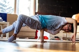 A person is performing a breakdance move in a living room. They are balancing on one hand with their body parallel to the floor. The room has a couch, a table, and visible daylight coming through a window.