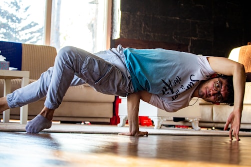 A person is performing a breakdance move in a living room. They are balancing on one hand with their body parallel to the floor. The room has a couch, a table, and visible daylight coming through a window.