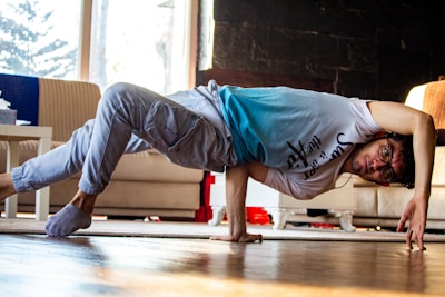 A person is performing a breakdance move in a living room. They are balancing on one hand with their body parallel to the floor. The room has a couch, a table, and visible daylight coming through a window.