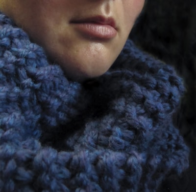 A close-up view of a person's lower face partially wrapped in a thick, knitted blue scarf. The texture of the scarf is prominently visible, with intricate yarn patterns.
