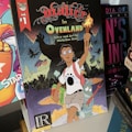 A comic book cover featuring a character with glasses holding a torch, accompanied by a creature, set in a fantastical landscape with flying creatures against a colorful backdrop. The title 'Malice in Ovenland' is prominently displayed with artistic lettering.
