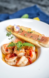 A beautifully presented dish of shrimp cooked in a flavorful sauce with herbs and red chilis, garnished with a sprig of fresh mint. Beside the shrimp, there is a piece of toasted bread topped with thin slices of ham and green herbs.