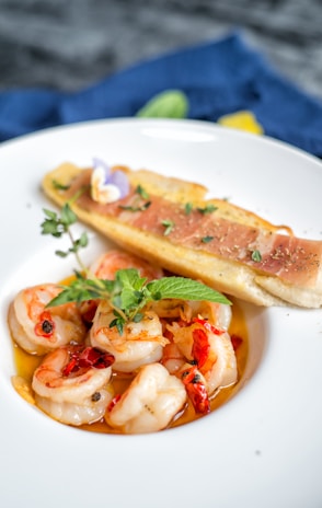 A beautifully presented dish of shrimp cooked in a flavorful sauce with herbs and red chilis, garnished with a sprig of fresh mint. Beside the shrimp, there is a piece of toasted bread topped with thin slices of ham and green herbs.