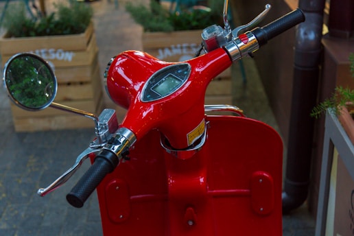 A close-up view of a bright red scooter handlebar with a round mirror and a small digital display. The handlebars have black rubber grips, and there is a chrome side view mirror to the left. In the background, wooden planters with greenery are partially visible.