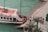 A vintage-style boat docked at a waterfront with several people walking along the pier. The boat has a red deck with white railings and life buoys attached. The water in the river is greenish, and there are palm trees near the walkway creating a serene environment.