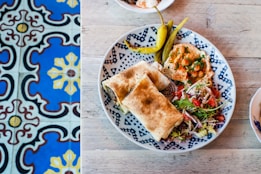 A neatly arranged plate with a variety of foods including wraps, hummus garnished with spices, pickled peppers, and a fresh salad with greens, tomatoes, and onions. The plate with a blue geometric pattern sits on a rustic wooden table. To the left, there are vividly colored tiles in blue, yellow, and maroon with intricate designs.