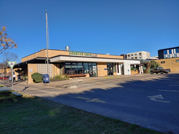A café situated on a corner, with visible signage indicating it offers breakfast, lunch, and dinner. Large windows display an open daily sign. There's an adjacent parking lot with a single vehicle. The sky is clear with bright sunlight casting shadows on the pavement. In the foreground, there are trees and a patch of grass along with a post office mailbox and a pedestrian sign.
