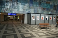 An indoor space with a tiled floor and a large wall featuring a mosaic pattern. Several advertising posters are displayed on panels along the wall. There is a wooden bench in the foreground and an electronic display with travel information above a corridor leading further into the building.
