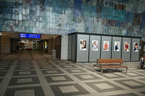 An indoor space with a tiled floor and a large wall featuring a mosaic pattern. Several advertising posters are displayed on panels along the wall. There is a wooden bench in the foreground and an electronic display with travel information above a corridor leading further into the building.