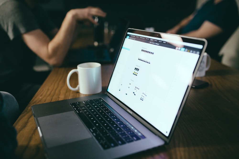 macbook pro beside white ceramic mug on brown wooden table