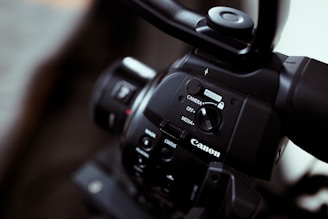 Close-up of a Canon video camera focusing on the control panel with buttons and switches, showing the brand logo prominently. The imaging device has a sleek black finish, and the lens is partially visible.