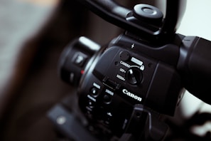 Close-up of a Canon video camera focusing on the control panel with buttons and switches, showing the brand logo prominently. The imaging device has a sleek black finish, and the lens is partially visible.
