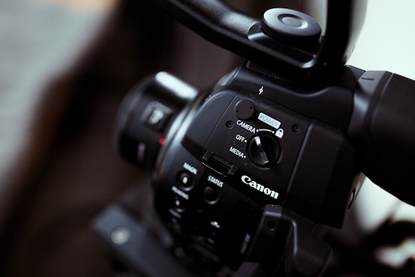 Close-up of a Canon video camera focusing on the control panel with buttons and switches, showing the brand logo prominently. The imaging device has a sleek black finish, and the lens is partially visible.