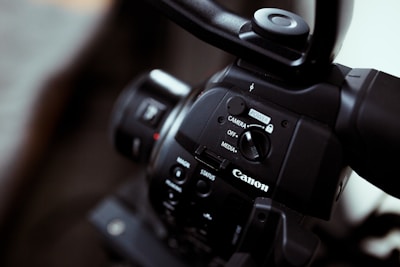 Close-up of a Canon video camera focusing on the control panel with buttons and switches, showing the brand logo prominently. The imaging device has a sleek black finish, and the lens is partially visible.