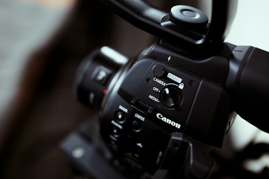 Close-up of a Canon video camera focusing on the control panel with buttons and switches, showing the brand logo prominently. The imaging device has a sleek black finish, and the lens is partially visible.