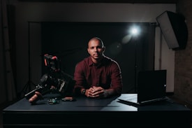 A person sits at a table with a serious expression, surrounded by filmmaking equipment such as a professional camera and a laptop. The setting is dimly lit, with a bright spotlight in the background that creates dramatic shadows.