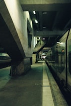 A train platform with a dark, industrial atmosphere. The concrete structures form geometric patterns above, while a train is positioned on the right. The platform is mostly empty, with dim lighting accentuating shadows.