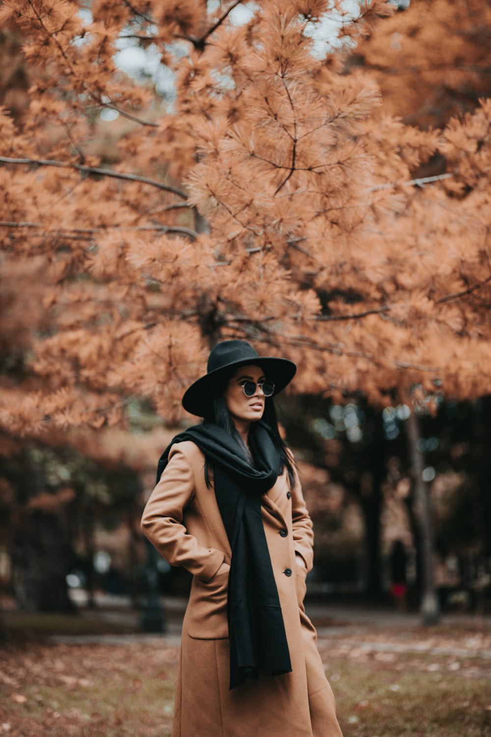 낮 동안 갈색 잎 나무 근처에 서 있는 갈색 코트를 입은 여자
