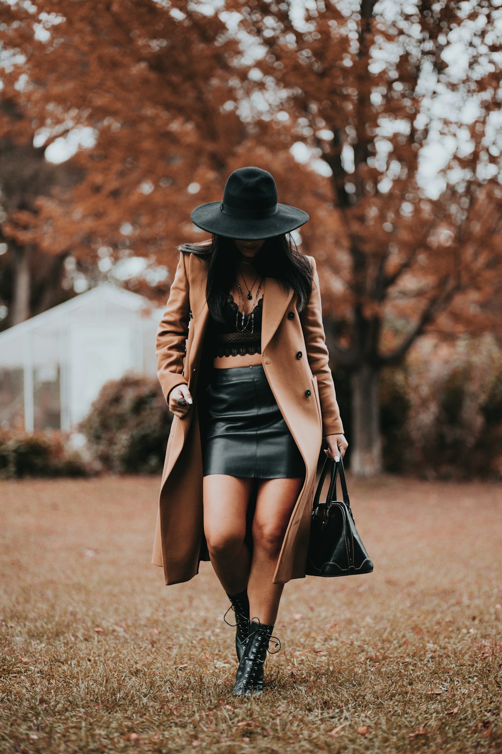갈색 코트와 검은 치마를 입은 여자가 검은 모자를 쓰고 갈색 잔디밭에 서 있다