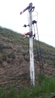 A vintage railway semaphore signal stands on a grassy embankment. The signal post is weathered, with peeling white paint, and is equipped with ladders and metal components. The background consists of grassy hills and overcast sky.