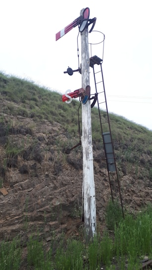 A vintage railway semaphore signal stands on a grassy embankment. The signal post is weathered, with peeling white paint, and is equipped with ladders and metal components. The background consists of grassy hills and overcast sky.