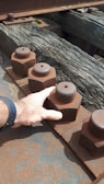 A close-up of a hand touching a large, rusted metal nut and bolt on an industrial structure. The bolts are lined up along a metal beam with a wooden surface beneath them.