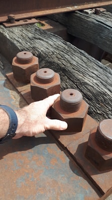 A close-up of a hand touching a large, rusted metal nut and bolt on an industrial structure. The bolts are lined up along a metal beam with a wooden surface beneath them.