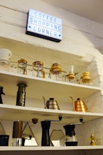 A brightly lit sign reading 'Coffee Shopping Corner' is mounted on a white brick wall above several shelves. The shelves are filled with various coffee-making equipment, including glass jars, a French press, hand grinders, and metal kettles in silver and copper hues. The overall arrangement of items is organized and neatly displayed.