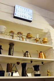 A brightly lit sign reading 'Coffee Shopping Corner' is mounted on a white brick wall above several shelves. The shelves are filled with various coffee-making equipment, including glass jars, a French press, hand grinders, and metal kettles in silver and copper hues. The overall arrangement of items is organized and neatly displayed.