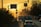 A city street during sunset with cars lined up and traffic signals visible. Street signs are pointing towards Sanayi Sitesi and Boğazkent. The scene is framed by silhouettes of palm trees and buildings, with a warm, golden hue dominating the sky.