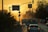 A city street during sunset with cars lined up and traffic signals visible. Street signs are pointing towards Sanayi Sitesi and Boğazkent. The scene is framed by silhouettes of palm trees and buildings, with a warm, golden hue dominating the sky.