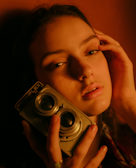 A person is holding a vintage twin-lens camera close to their face, with a soft and introspective expression. The lighting is warm and creates a moody, nostalgic atmosphere.