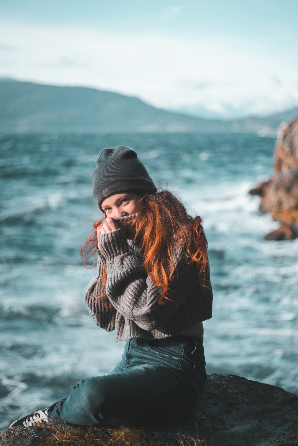 woman in black knit cap and black jacket standing on rock formation near body of water