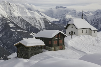 Snow-covered alpine landscape featuring two wooden cabins and a small church. The buildings are surrounded by thick layers of snow. Behind them, a dramatic range of snow-capped mountains extends into the distance, partially shrouded by clouds.