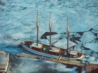 An aerial view of a large sailboat moored at a dock with a frozen, cracked ice surface surrounding it. The boat is a classic wooden schooner with three masts. The intricate patterns of the ice create a mosaic-like texture across the water.
