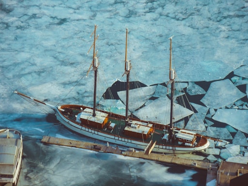 An aerial view of a large sailboat moored at a dock with a frozen, cracked ice surface surrounding it. The boat is a classic wooden schooner with three masts. The intricate patterns of the ice create a mosaic-like texture across the water.