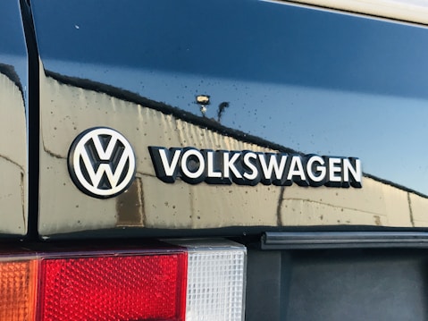 Close-up view of a Volkswagen logo and the name on a dark blue vehicle, with reflections of light on the wet surface and part of the taillight visible.