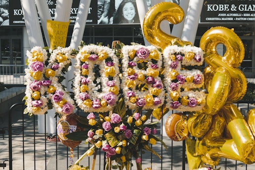 A floral tribute with the word 'KOBE' spelled out in flowers, featuring white, yellow, and pink blooms. Gold balloons are positioned around the arrangement. A sign with the message 'Rest in Paradise Kobe' is visible, surrounded by other floral arrangements and Los Angeles Lakers balloons.
