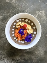 A beautifully arranged smoothie bowl with sliced bananas, chopped papaya, and a single red raspberry on top. The bowl also features vibrant purple flowers and white dollops around the rim on a creamy base.