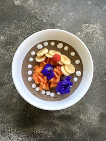A beautifully arranged smoothie bowl with sliced bananas, chopped papaya, and a single red raspberry on top. The bowl also features vibrant purple flowers and white dollops around the rim on a creamy base.