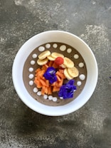 A beautifully arranged smoothie bowl with sliced bananas, chopped papaya, and a single red raspberry on top. The bowl also features vibrant purple flowers and white dollops around the rim on a creamy base.